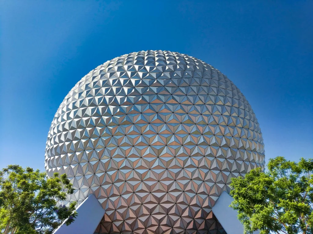 Cúpula geodésica metálica do Epcot na Disney Orlando com padrão de triângulos em tom prateado e azul, vista de baixo, acima de árvores verdes e céu azul