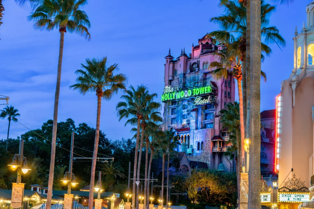 Torre do The Hollywood Tower Hotel em Hollywood Studios com palmeiras altas ao entardecer, luzes decorativas e céu azul ao fundo.