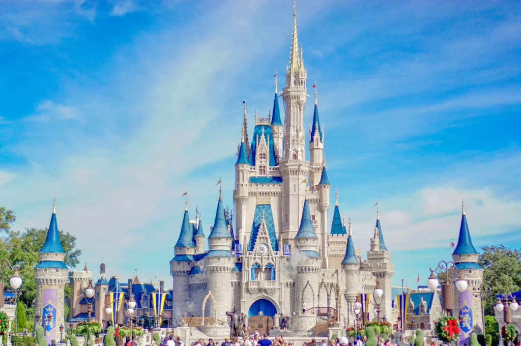 Castelo da Cinderela no Magic Kingdom com torres azuis e brancas, fachada imponente e entrada principal em destaque diante de céu azul e multidão na frente, em um dia ensolarado.