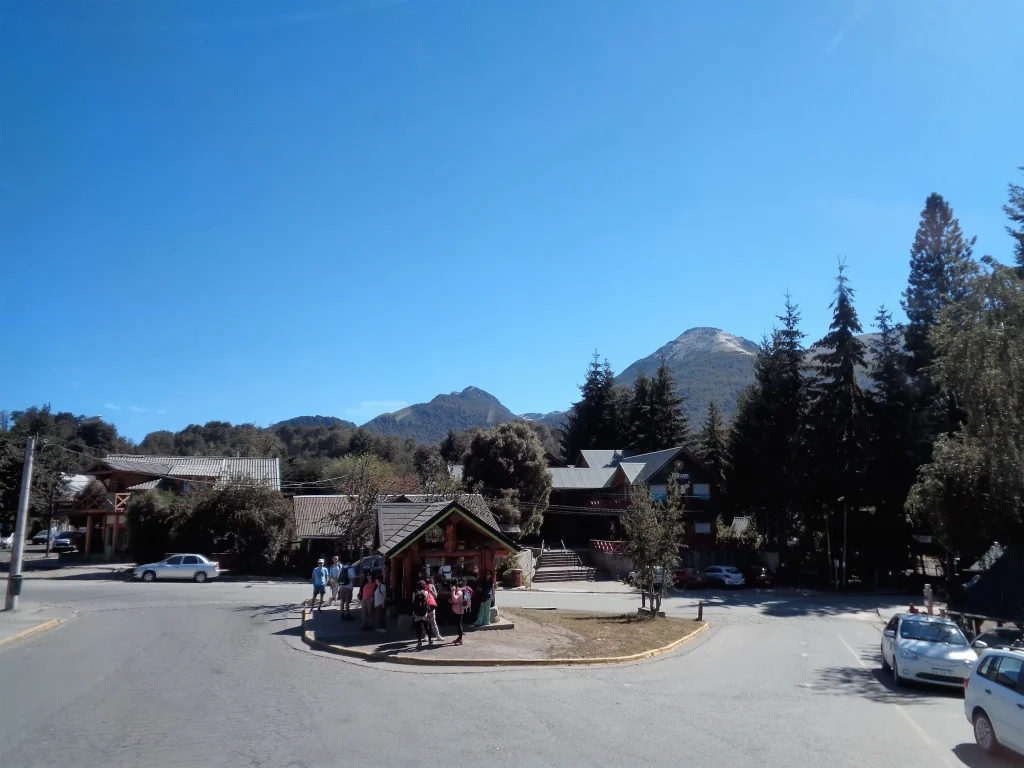 Foto de Villa La Angostura, con pequeña casa al centro, pineros a la derecha y la gente alrededor.