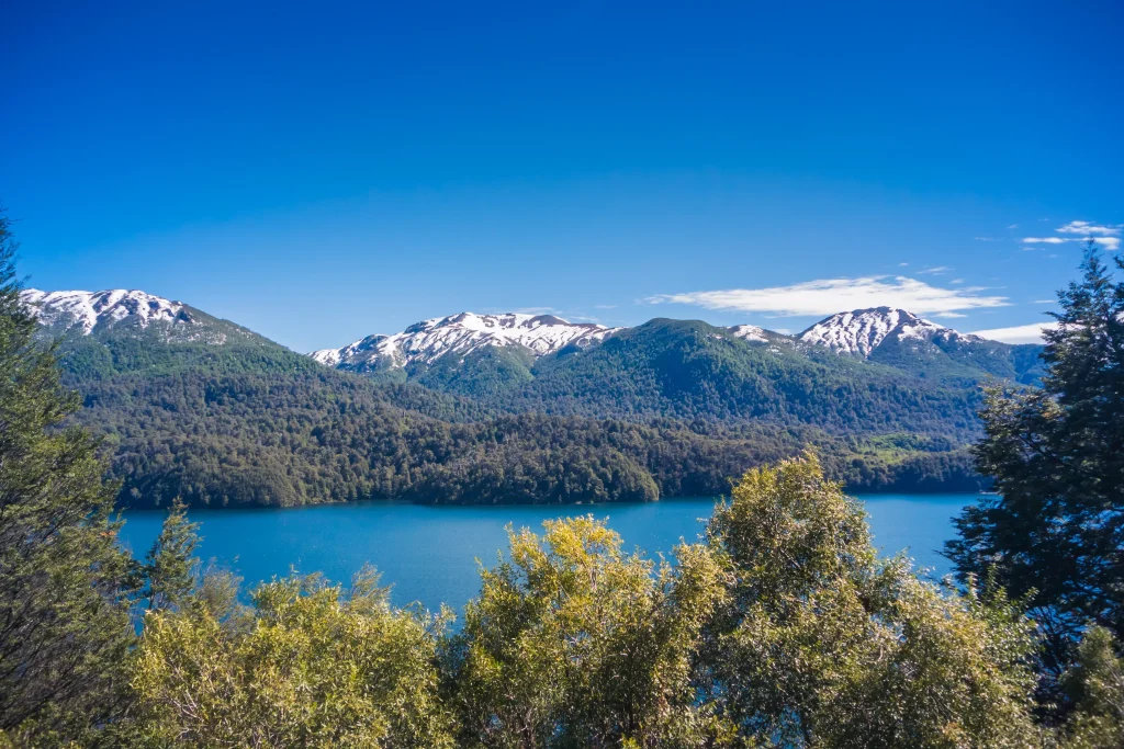 Vista desde arriba del Lago Villarino, una de las maravillas que vas a encontrar en el camino de los 7 lagos en auto. La foto tiene el cielo azul, montañas al fondo y árboles en frente.