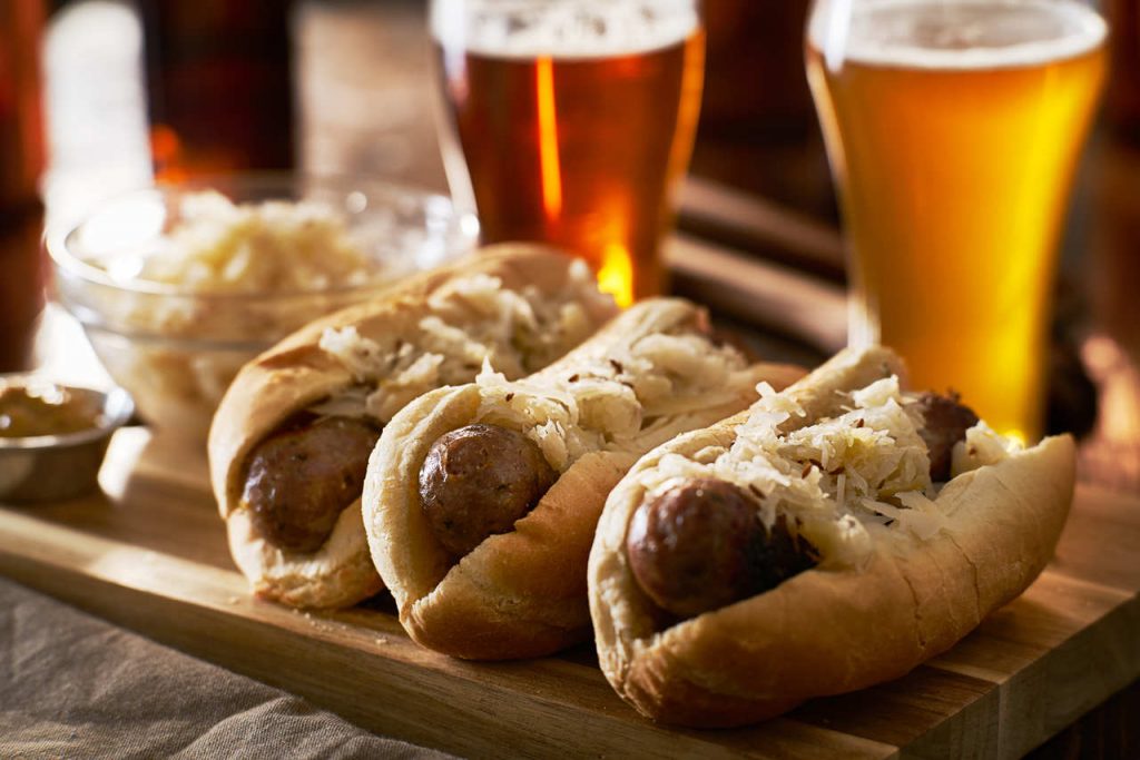 Três pães com salsicha alemã e copos de cerveja ao fundo.