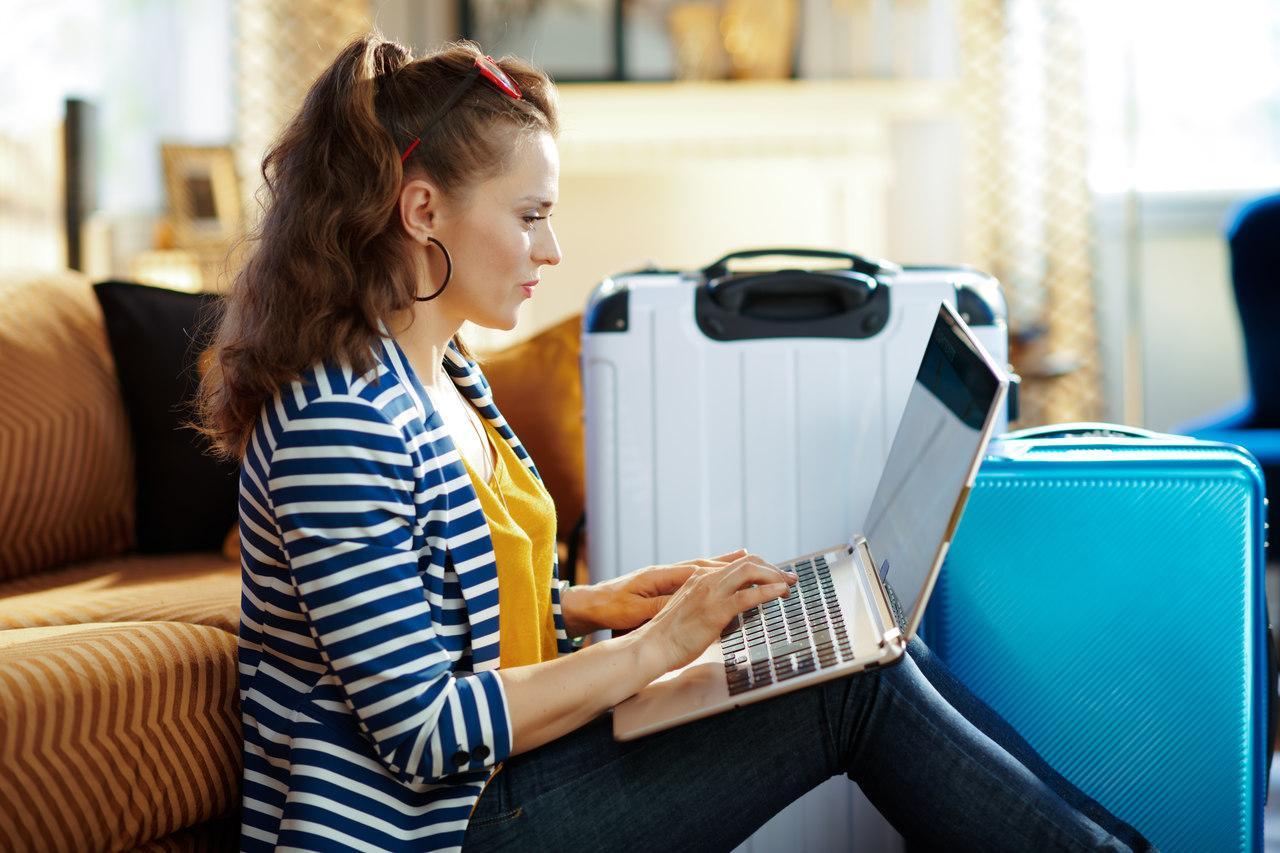 Mulher com um notebook no colo, mexendo no computador, com malas de viagem no fundo do ambiente
