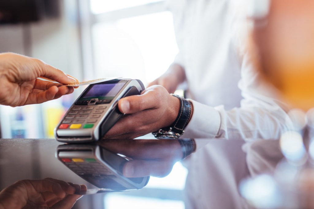 Imagem focada em uma mão masculina segurando um cartão para passar em uma máquina de cartão de crédito