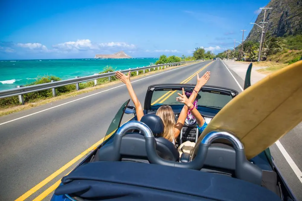 Foto de dos mujeres con los brazos arriba en un auto en una carretera cerca del mar.