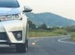 Parte frontal de um carro branco na estrada, com faróis e grade visíveis, seguindo pela via em meio a paisagem de montanhas ao fundo.