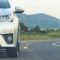 Parte frontal de um carro branco na estrada, com faróis e grade visíveis, seguindo pela via em meio a paisagem de montanhas ao fundo.