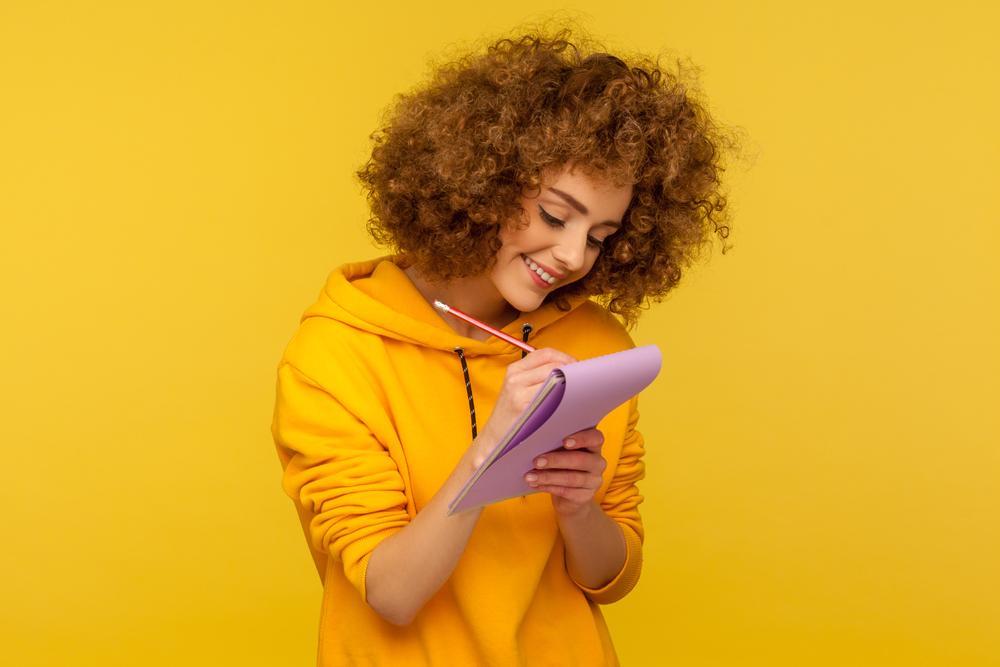 Mulher usando roupa amarela está com um bloco de notas em mãos para anotar tudo que precisa para economizar na viagem 