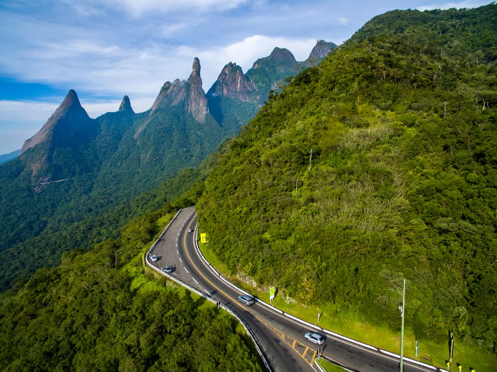 Foto de estrada com uma imagem bela da natureza, floresta bem verde e diversos picos montanhosos ao fundo.