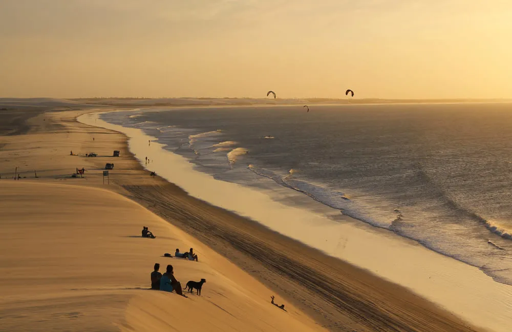 Praia de Fortaleza, Ceará, com dunas de areia, um mar com ondas e o entardecer no céu.