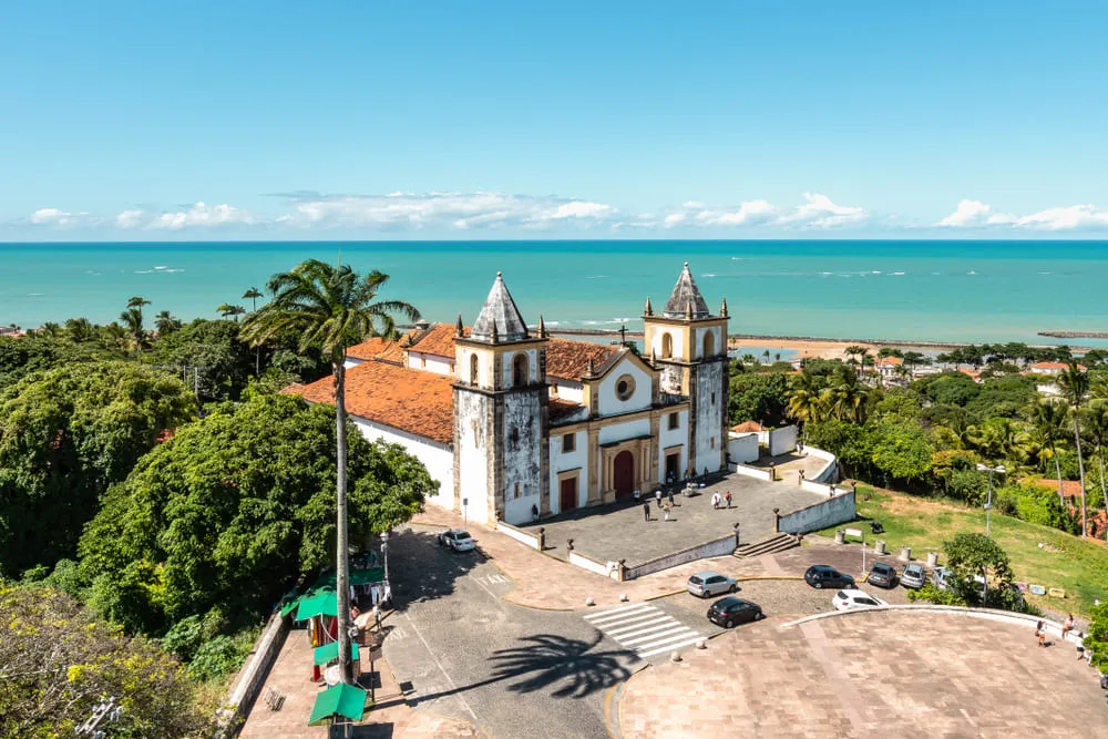 Foto de Olinda, em Pernambuco, com a praia ao fundo, o céu azul, coqueiros, árvores bem verdes e uma igreja antiga ao centro.