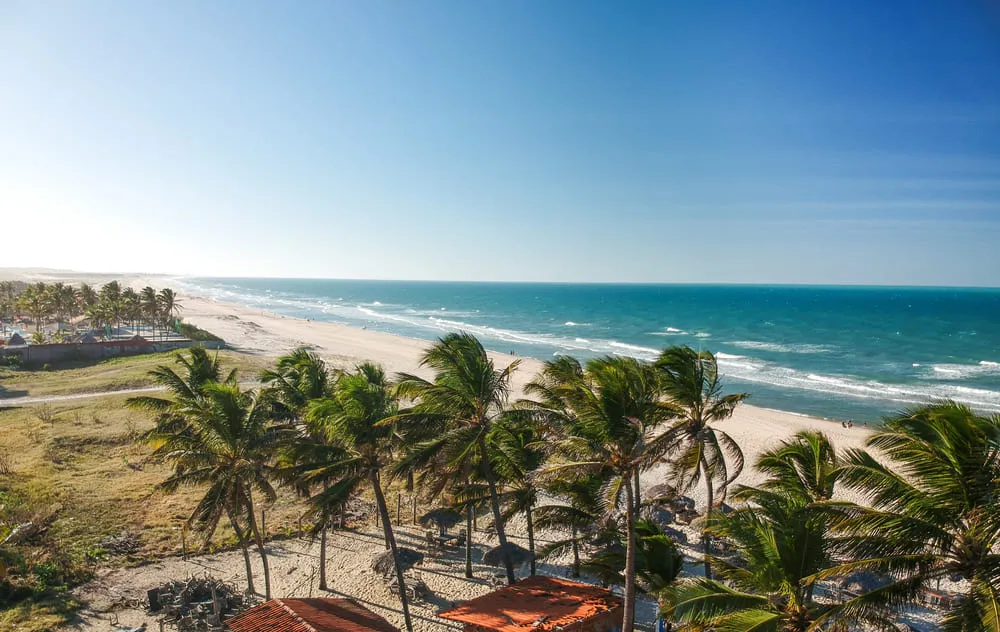 Foto de Fortaleza, Ceará, com a praia de mar azul e céu azul, repleta de coqueiros verdes e uma faixa de areia bem grande.