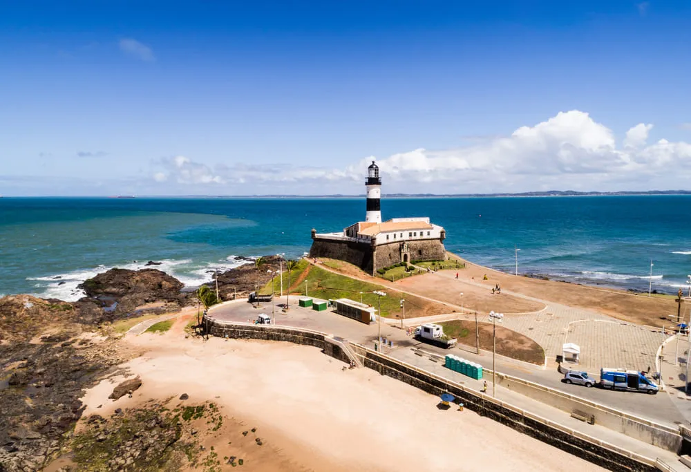 A Praia do Farol fica em Salvador, na Bahia. O Farol fica ao centro da imagem, o céu está bem azul e há pedras e areia na imagem.