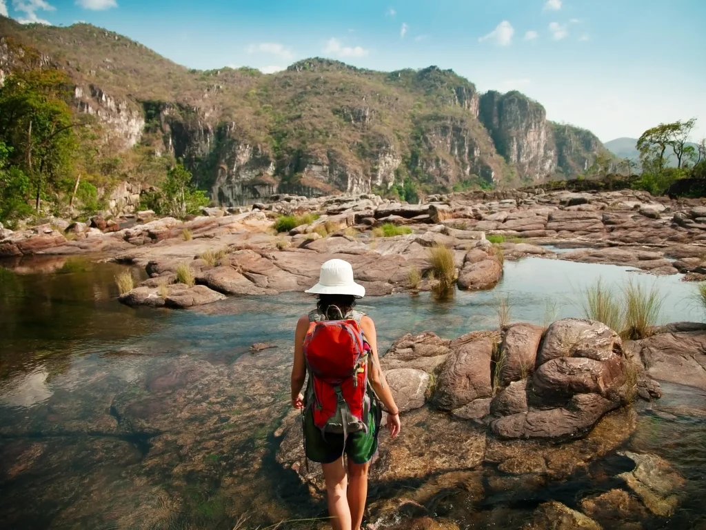 Chapada dos Veadeiros, em Goiás. Imagem mostra uma mulher, de costas, andando pelo riacho entre formações rochosas. Ao fundo, montanhas e muitas árvores.