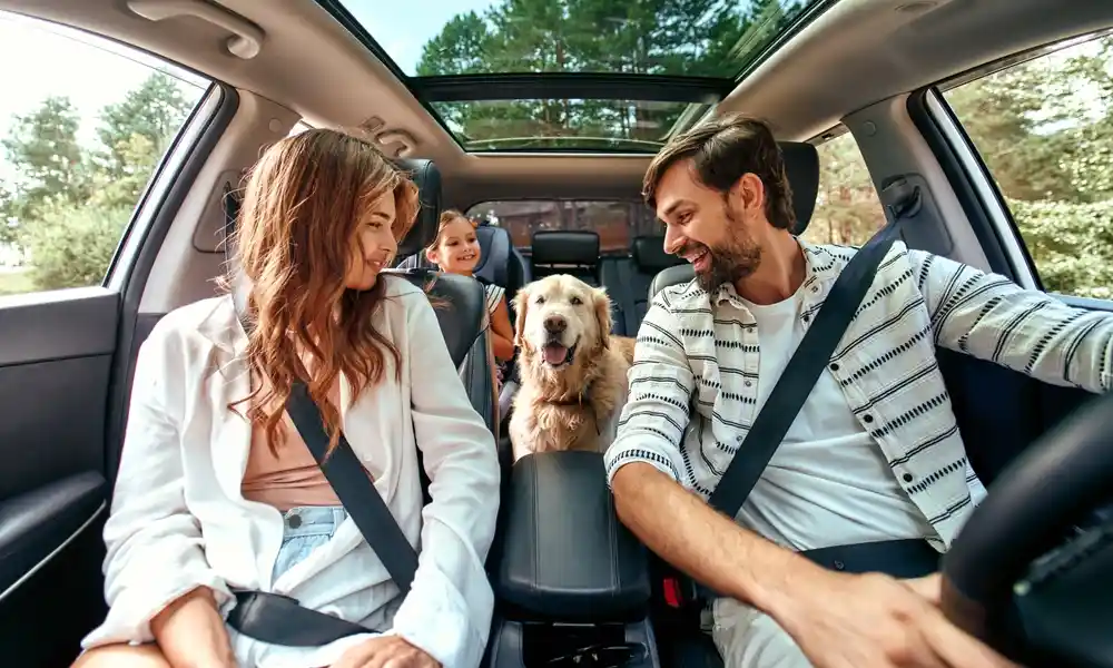 Casal no carro com cachorro labrador no meio e a filha sentada na cadeirinha na parte de trás do carro alugado