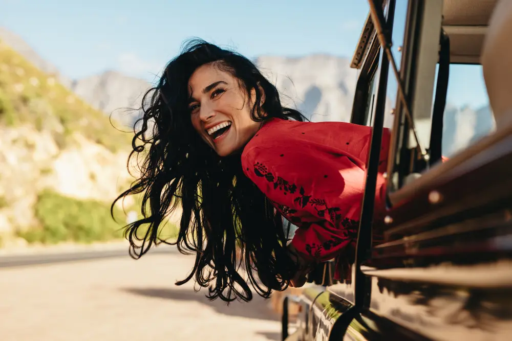 Mulher com cabelos pretos e camisa vermelha sorrindo com o corpo para fora da janela de um carro parado