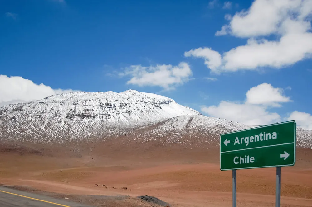 Foto da estrada com paisagem de montanhas nevadas ao fundo e a placa que indica Argentina e Chile para direções opostas.