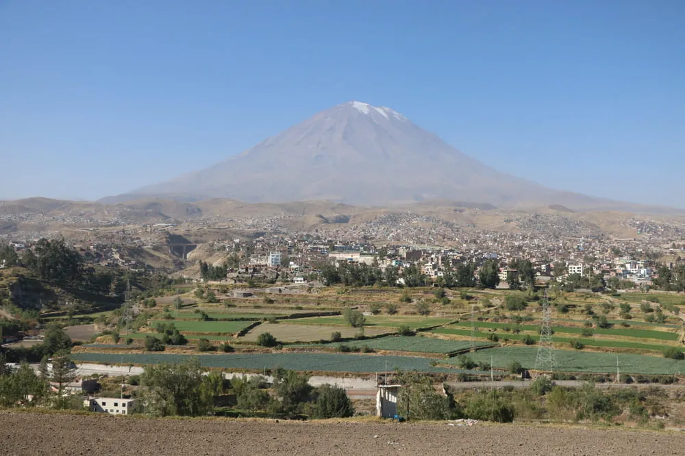 Imagem incrível do vulcão do Peru ao fundo, com seu pico nevado e a cidade abaixo, com muita vegetação preenchendo a paisagem.