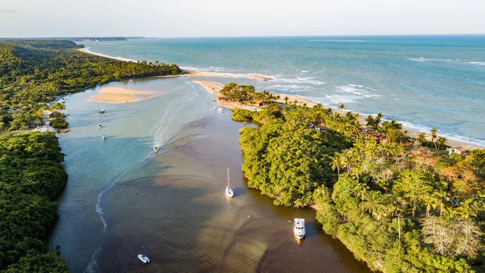 Foto de Caraíva, Bahia. Na foto é possível ver o encontro do mar com o rio, alguns barcos e muito verde da natureza.