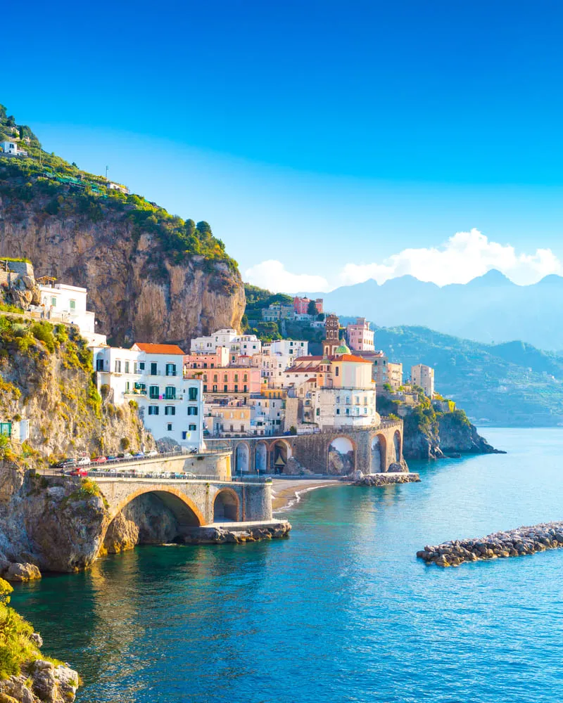 Foto do Sul da Itália, com suas casinhas coloridas e enfileiradas com o mar ao lado. O céu é azul e ao fundo tem montanhas.