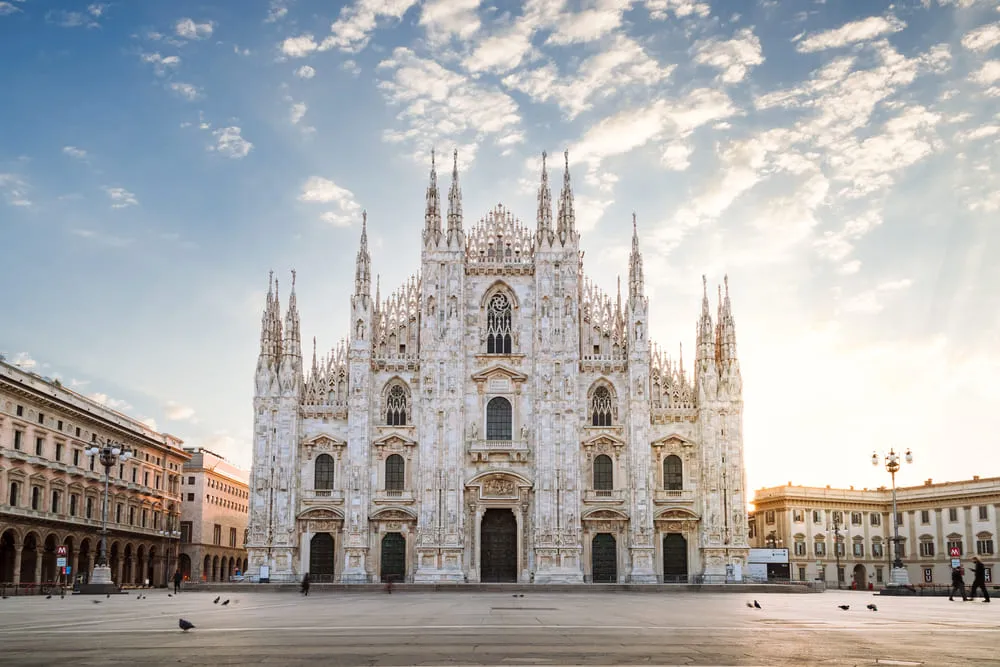 Foto do Duomo de Milão e toda sua arquitetura gótica. O céu está azul e há pássaros na frente do Duomo.