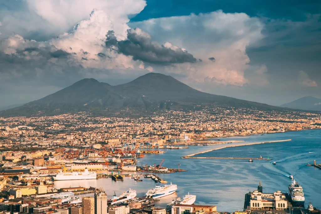 Foto aérea mostra a cidade de Nápoles, na Itália, com o mar banhando a cidade, os morros ao fundo e o céu azul.