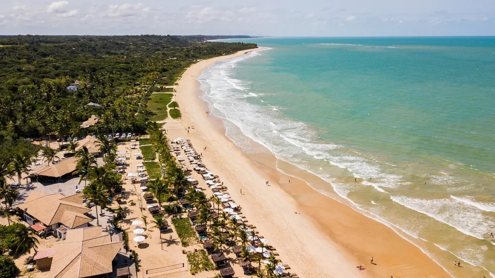 Foto de Porto Seguro, na Bahia. A foto apresenta uma vista aérea repleta de verde e o azul do mar. A praia apresenta uma longa faixa de areia.