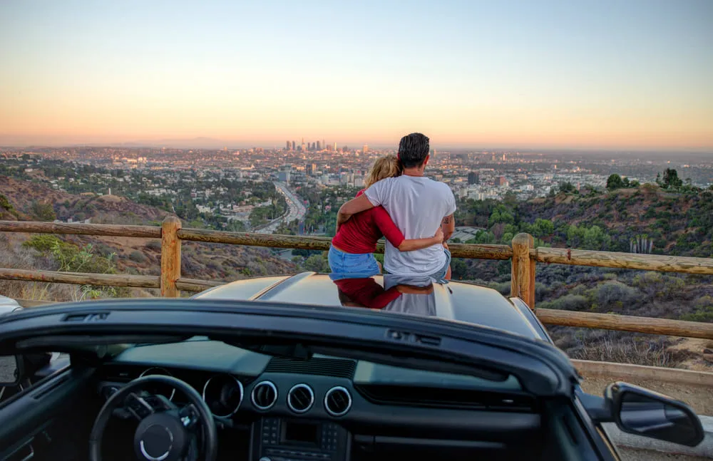 Foto de um casal em cima do capô do carro vendo Los Angeles de cima. A vista é espetacular das clinas, prédios e natureza.