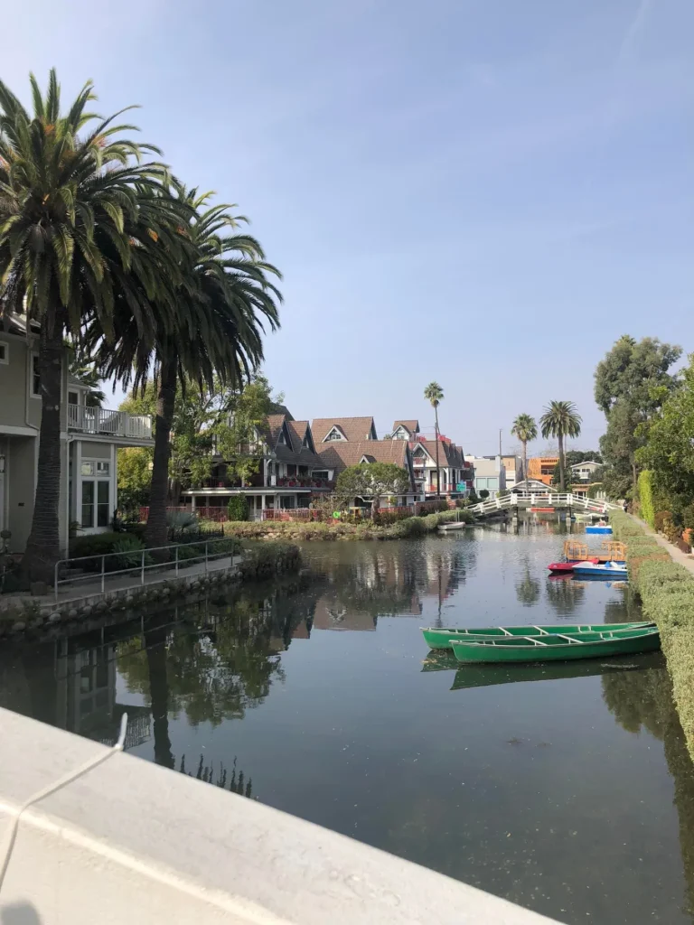 Foto dos Canais de Venice, com as casas ao redor do rio que passa. Alguns barcos estão parados nas extremidades. O céu está bem azul e tem palmeiras ao redor.