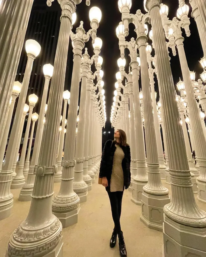 Foto em frente a infinidade de postes de luz famosos do o LACMA (Los Angeles County Museum of Art). Há uma mulher em frente e está de noite.