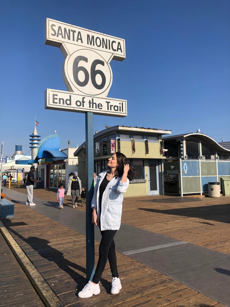 Foto de uma mulher em frente a placa que indica "Santa Monica 66 - End Of The Trail" que fica no Píer de Santa Mônica, na Califórnia.