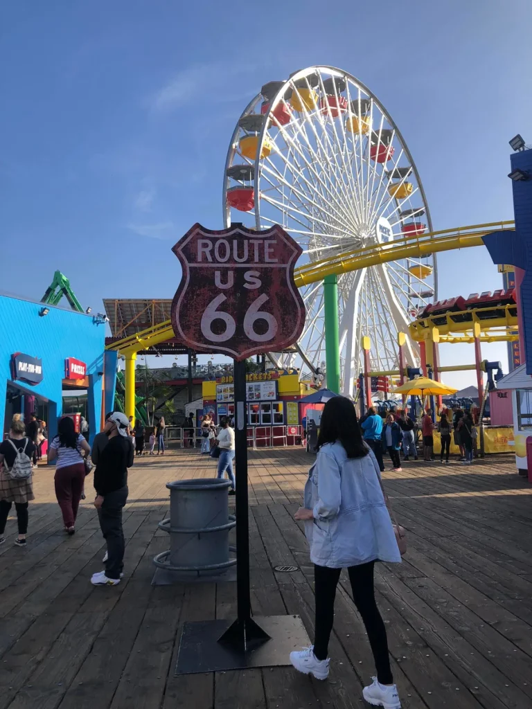 Foto do parque de diversões Pacific Park, no Píer de Santa Mônica, em Los Angeles. O dia está azul e tem uma mulher em frente à placa que indica Route 66.