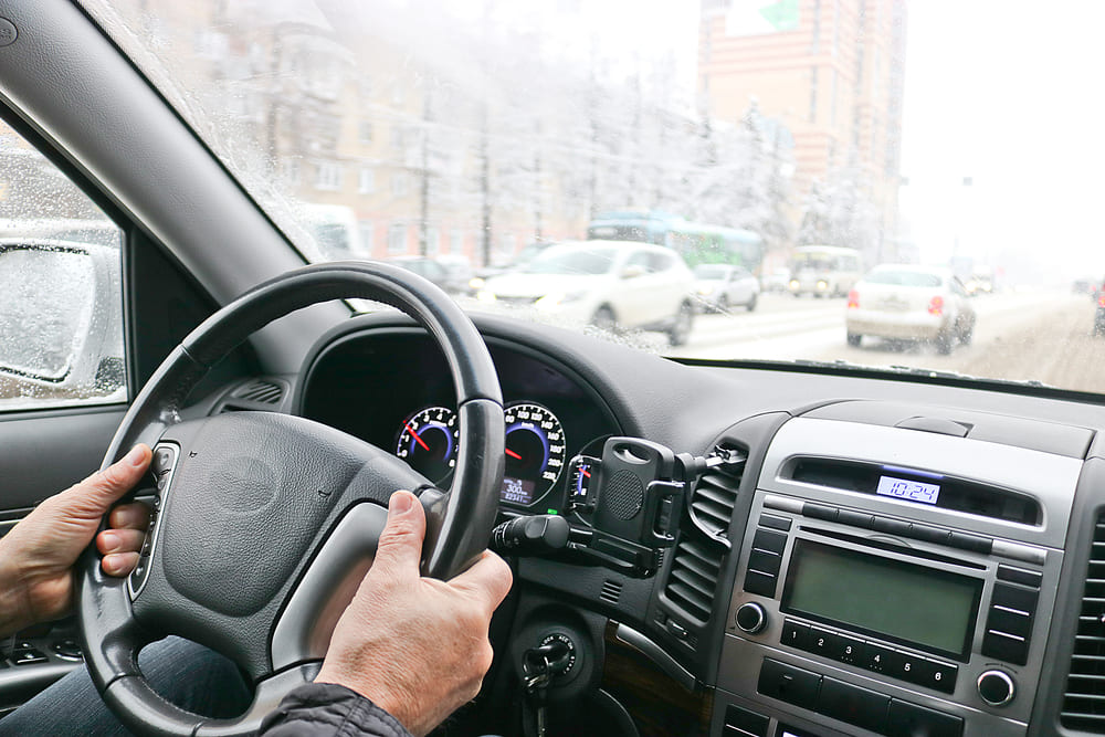 Foto do interior de um carro, com um homem com as mãos no volante. No exterior é possível observar a cidade e o trânsito de carros.