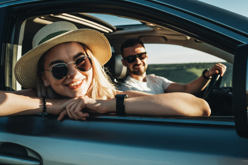 Foto de um casal sorridente dentro do carro. A mulher está com óculos de sol e chapéu.