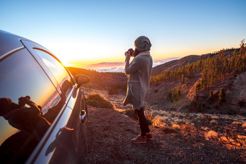 Mulher tirando foto com câmera profissional da paisagem espetacular da natureza. O céu está bem azul e o carro está estacionado ao lado da mulher.