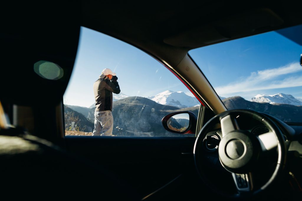Foto do interior de um carro com um viajante ao lado de fora, registrando uma foto com sua câmera profissional. O dia está azul e os picos nevados estão ao fundo.
