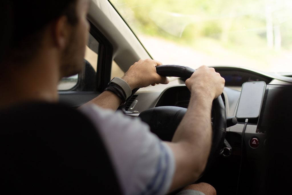 Foto de uma pessoa dirigindo um carro, com as mãos no volante e o dia claro no exterior.