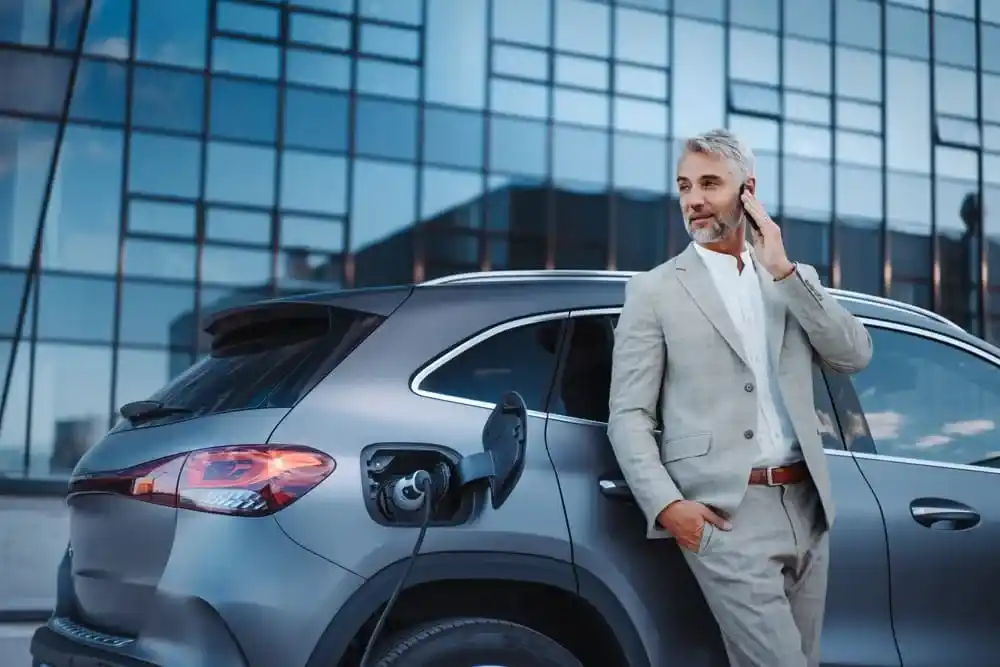Foto de um senhor em frente a um carro elétrico, ele está no celular enquanto aguarda o carro ser carregado eletricamente completamente.