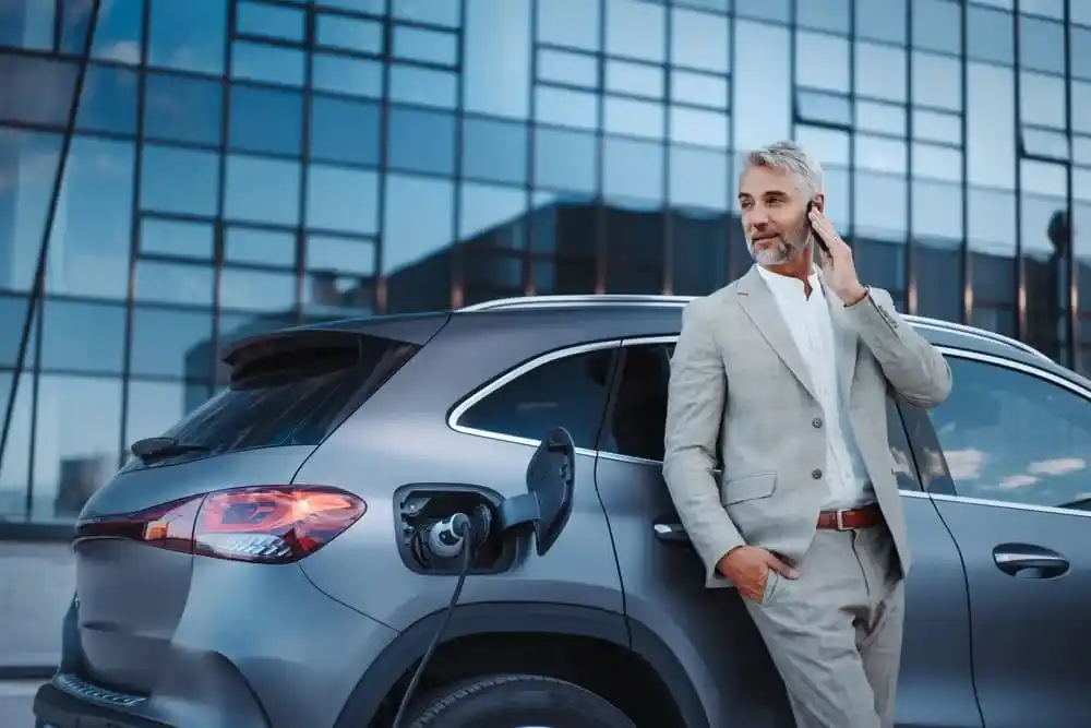 Homem esperando a recarga do seu carro elétrico ser finalizada. Atrás tem um prédio comercial.