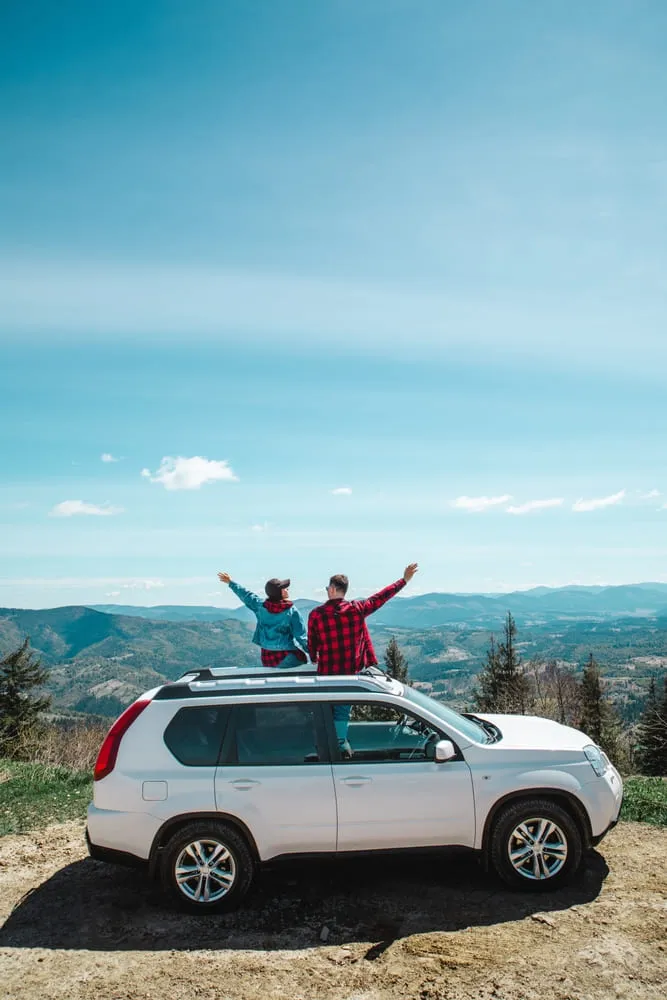 Foto de carro SUV com um casal sentado em cima. O carro é branco, 4 portas. O céu está azul e é possível ver o horizonte de natureza.
