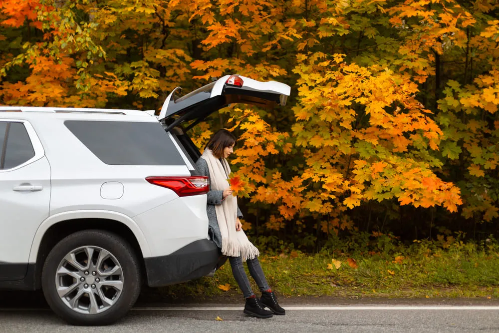 Foto de uma mulher sentada no porta malas do carro, que está com a porta levantada. O carro é um SUV branco.