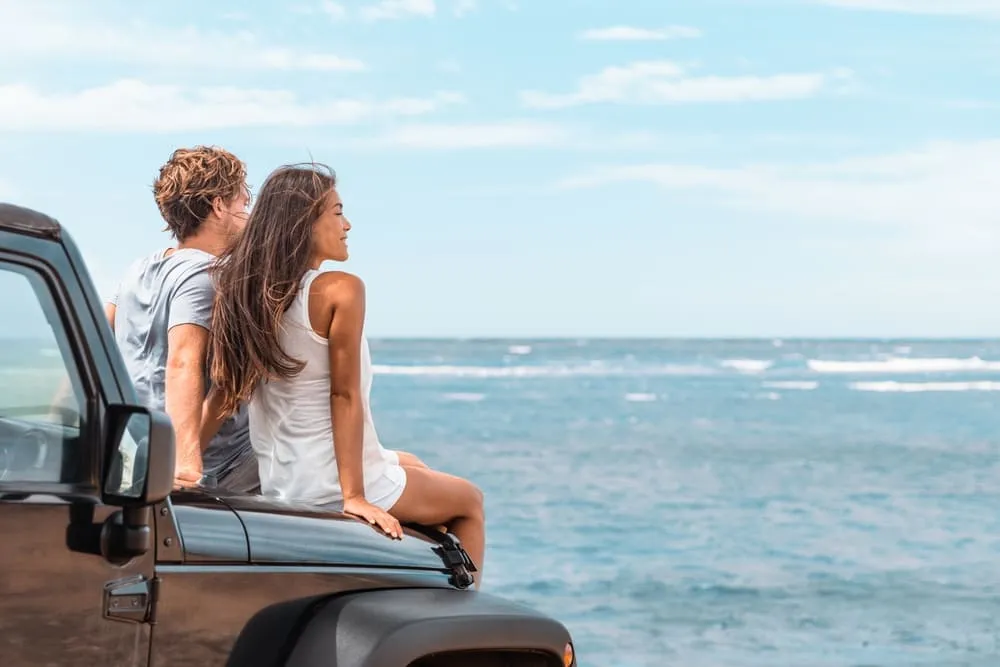 Um casal em cima do capô do carro, observando o horizonte e o mar ao fundo. Está calor e o céu está bem azul.