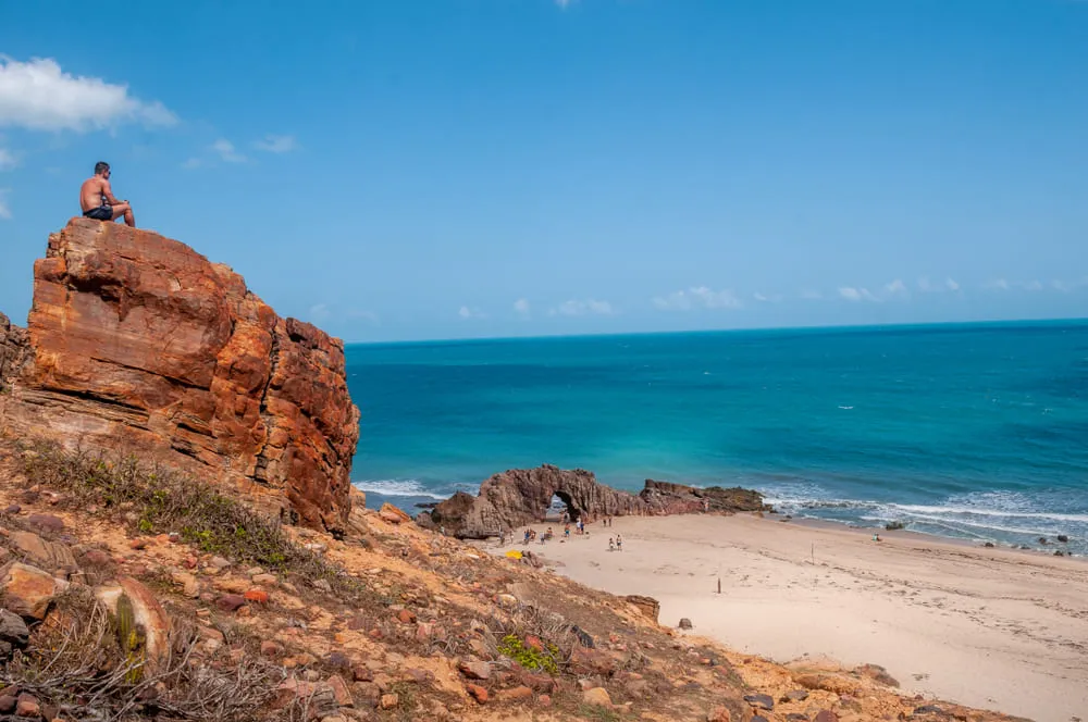 Foto da Pedra Furada em Jericoacoara, região Nordeste do Brasil. O mar e o céu estão na cor azul. Há um homem sentado em cima de uma pedra observando a paisagem. Muitas pessoas fazem foto na Pedra Furada.