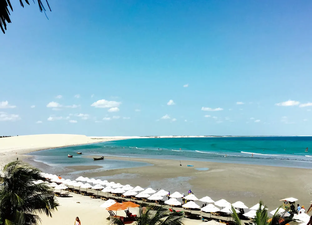 Foto de uma das praias de Jericoacoara, região Nordeste do Brasil. É possível ver uma infinidade de guarda-sol na beira da praia, todos brancos e padronizados. O mar é bem azul e é possível ver as lagoas formadas pelo mar. No fundo, vemos as dunas de areia bem branquinha.