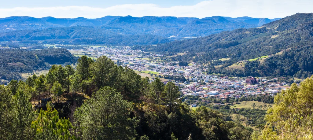 Foto aérea de Urubici na Serra Catarinense. É possível ver as montanhas impressionantes, o céu azul, a cidade ao fundo e a vegetação bem verde.