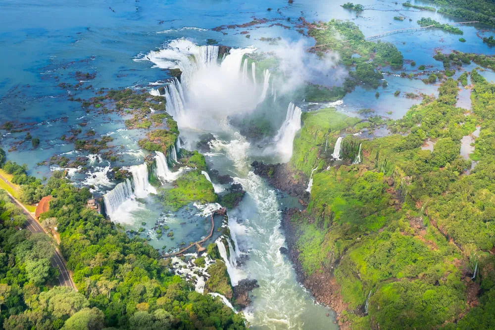 Foto aérea das Cataratas do Iguaçu no Parque Nacional do Iguaçu, em Foz do Iguaçu. É possível ver as quedas de água e a vegetação local. 