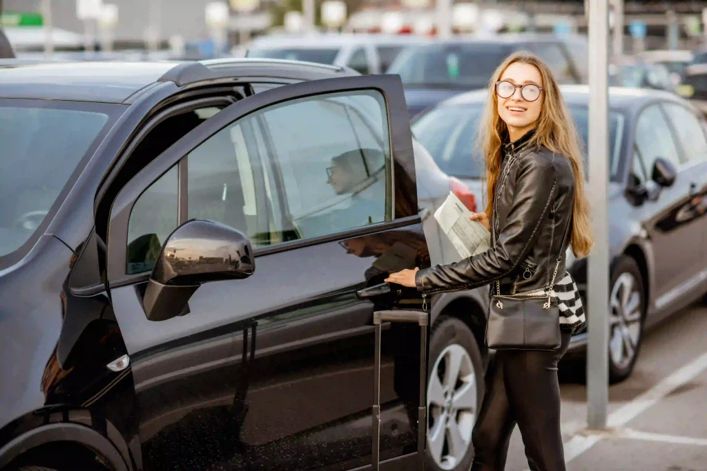 Mulher abrindo a porta de um carro preto, ela está com mala de viagem e sorrindo para foto. O carro está em um estacionamento.