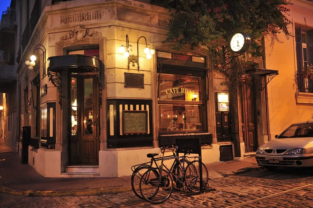 Foto do Café Rivas na cidade de Buenos Aires, na Argentina. A construção é super vintage, dando um ar mais sofisticado para a foto. A rua é de pedras, há um carro e duas bicicletas estacionados em frente.