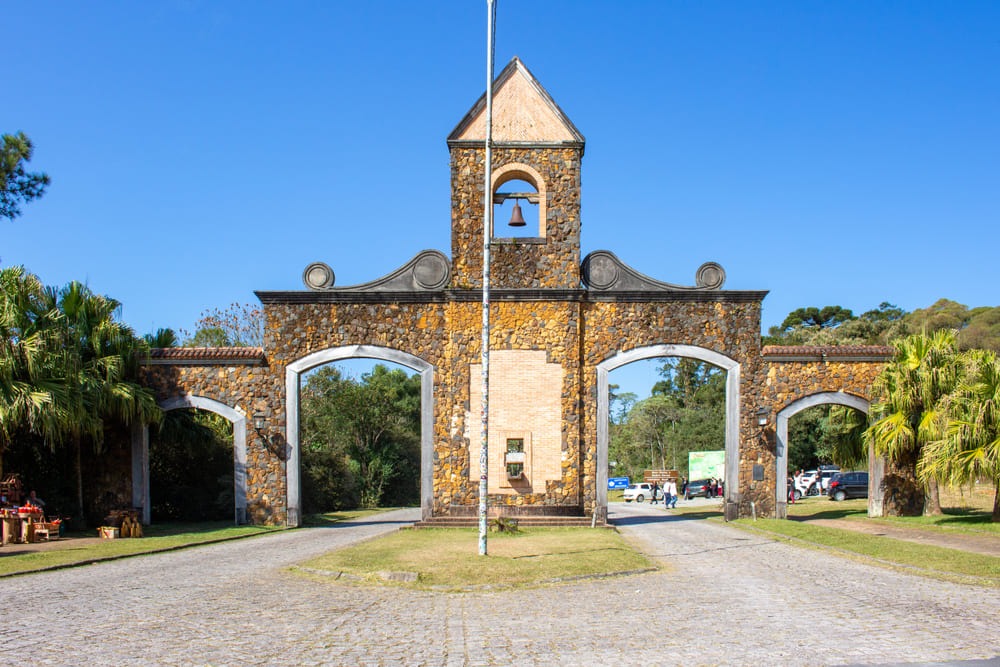 Foto do portal da Estrada da Graciosa que leva até Morretes, no Paraná. O portal é feito de pedras marrons e existe um sino ao centro e no topo.