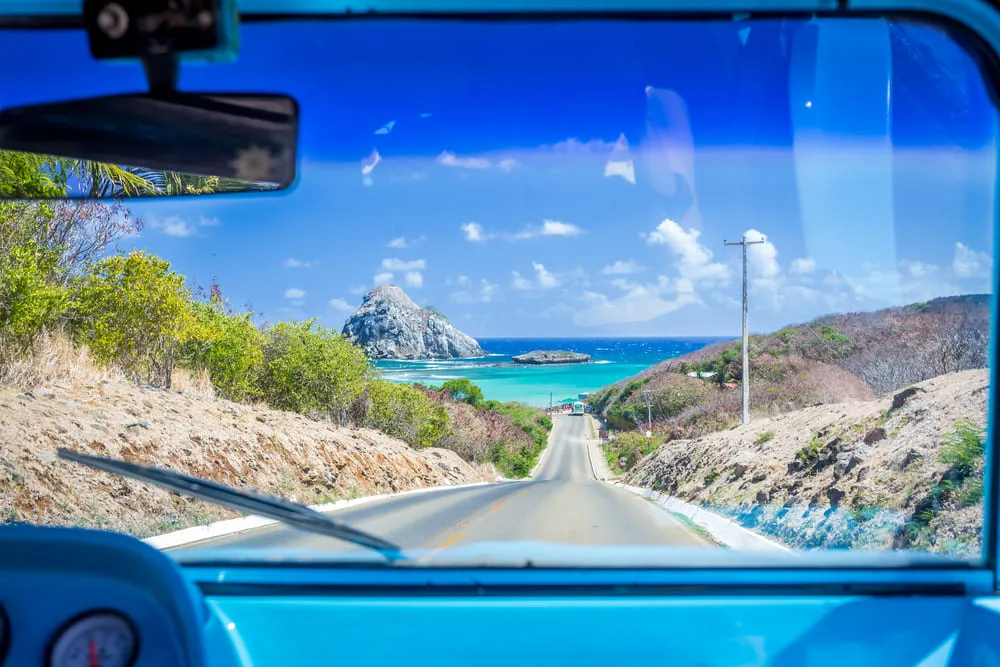 Foto da visão de dentro do carro para a praia de Fernando de Noronha. O céu e o mar são bem azuis. A estrada mostra a rua até chegar na areia.
